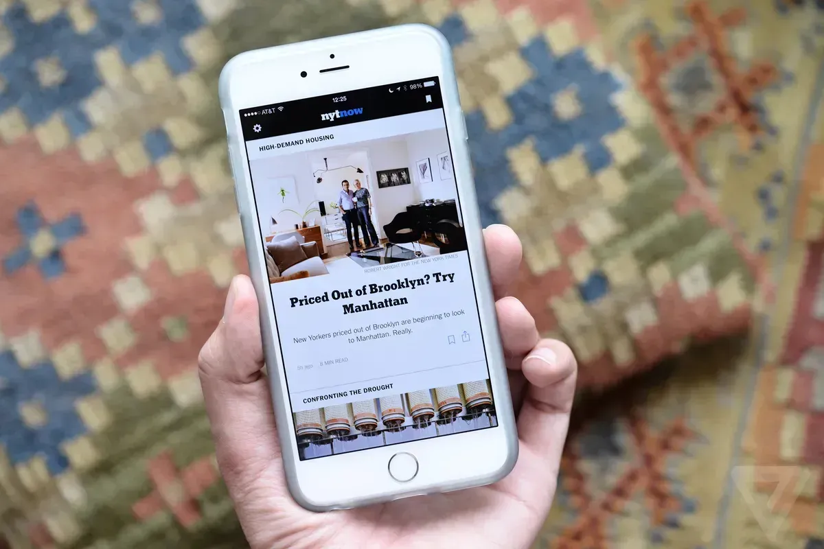 A hand holding a phone showing the NYT Now app